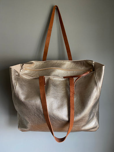 Large gold tote bag with zip top shown hanging from a hook on the wall. 