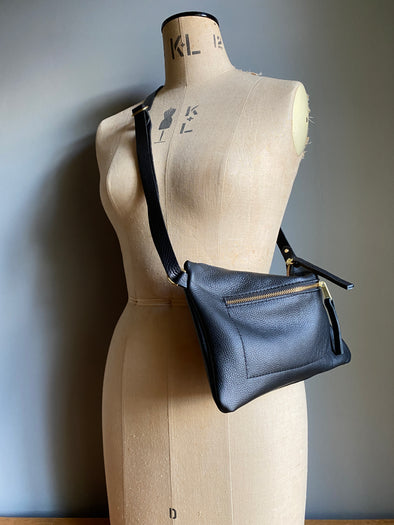 Crossbody handbag in black leather with zipped exterior pocket and crossbody strap by Ginger and Brown shown on a vintage tailor's dummy. 