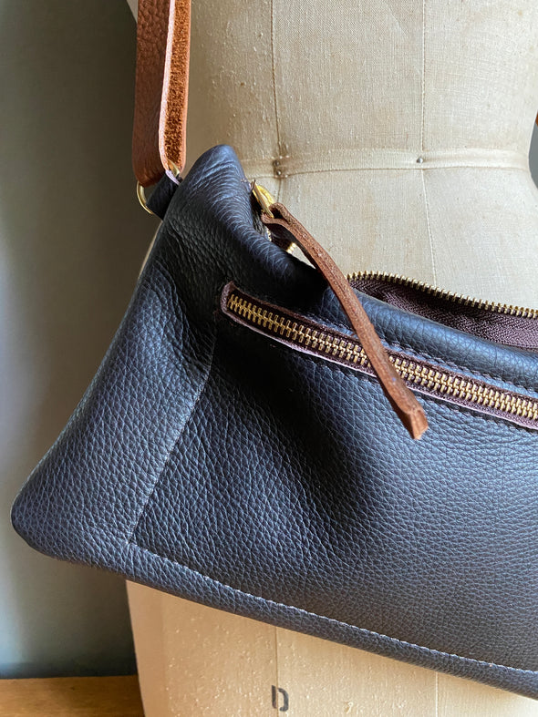 Crossbody handbag in dark brown leather with zipped exterior pocket and contrasting brown strap by Ginger and Brown shown on a table top.