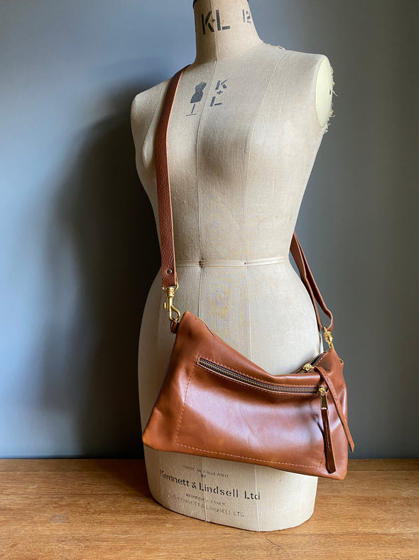 Crossbody handbag in tan brown leather with zipped exterior pocket and adjustable crossbody strap by Ginger and Brown shown on a vintage tailor's dummy. 