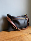 Crossbody handbag in dark brown leather with zipped exterior pocket and contrasting brown strap by Ginger and Brown shown sitting on a table top. 