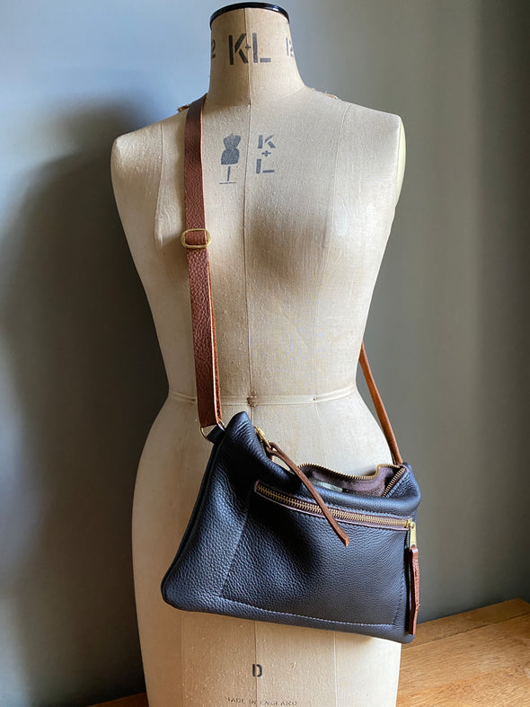 Crossbody handbag in dark brown leather with zipped exterior pocket and contrasting brown strap by Ginger and Brown shown sitting on a table top. 