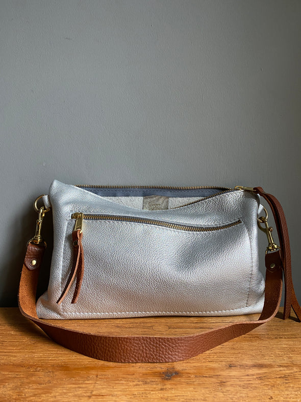 Crossbody handbag in silver leather with zipped exterior pocket and contrasting brown strap by Ginger and Brown shown sitting on a table top.