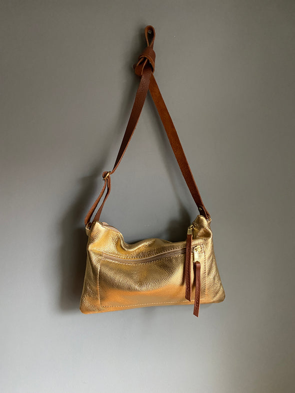 Gold leather crossbody handbag with zipped exterior pocket by Ginger and Brown shown hanging from a hook with the strap tied in a knot. 