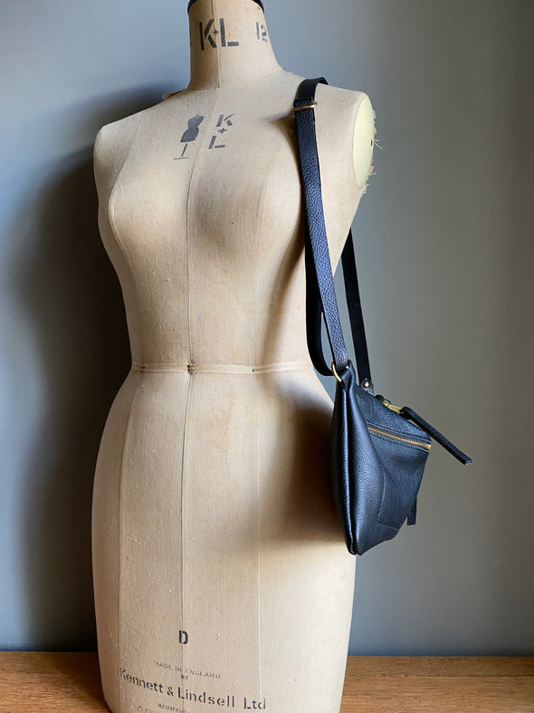 Crossbody handbag in black leather with zipped exterior pocket and adjustable crossbody strap by Ginger and Brown shown on a vintage tailor's dummy. 