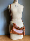 Crossbody handbag in tan brown leather with zipped exterior pocket and adjustable crossbody strap by Ginger and Brown shown on a vintage tailor's dummy. 