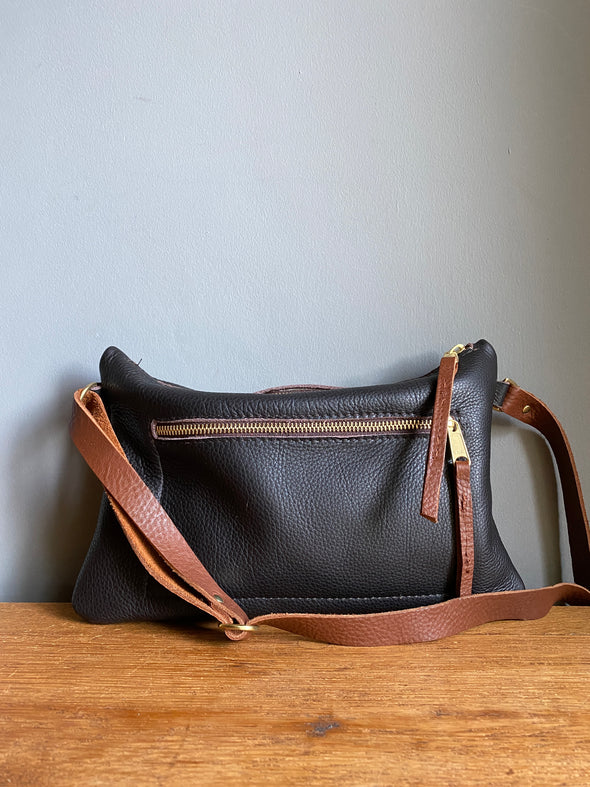 Crossbody handbag in dark brown leather with zipped exterior pocket and contrasting brown strap by Ginger and Brown shown sitting on a table top. 